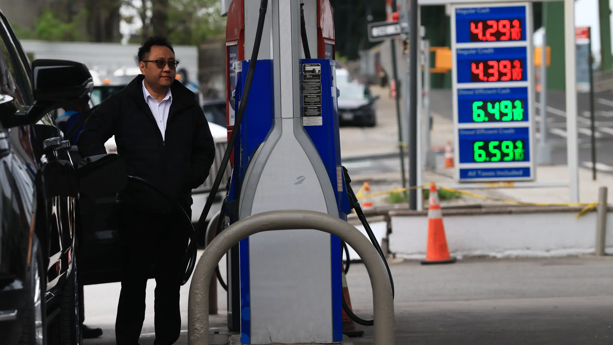 A person stands between a gas pump and a vehicle, with gas prices of $4.29 for a gallon of regular displayed in the background.