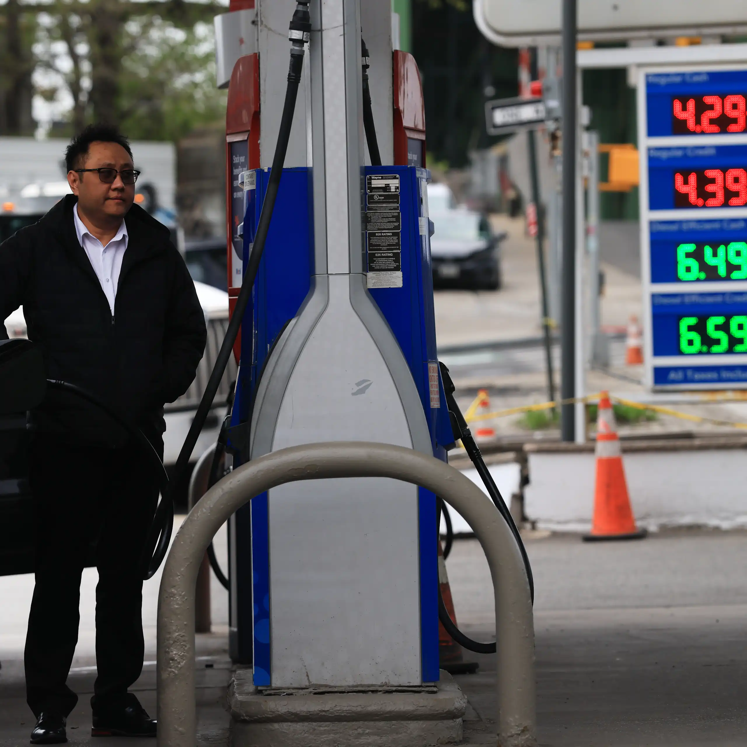 A person stands between a gas pump and a vehicle, with gas prices of $4.29 for a gallon of regular displayed in the background.