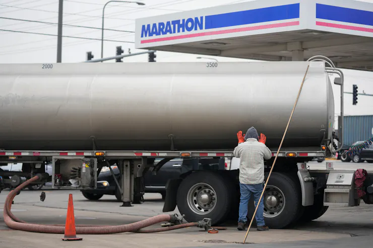 A person stands near a long metal truck in front of a gas station.