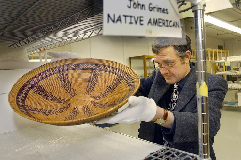 A man with white gloves picks up a woven brown bowl.