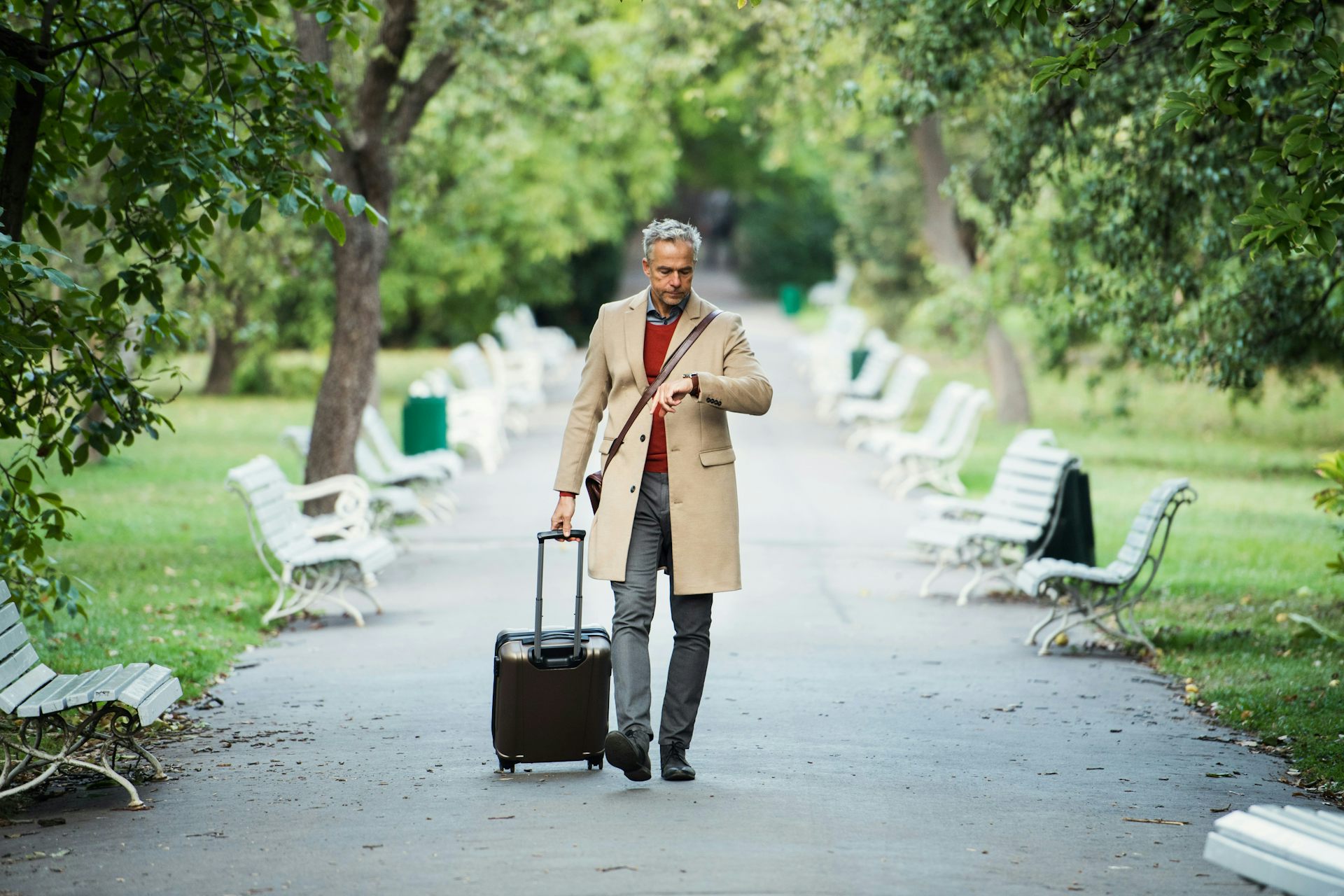 A grey-haired man pulls luggage through a city park while looking at his watch.