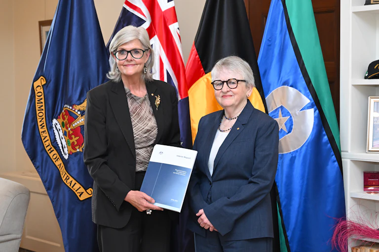 Two wmen with glasses stand in front of multiple flags and hold a blue and white document