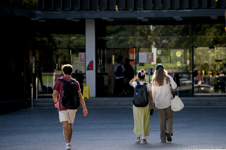 Three young people walk on university campus, heading towards the doors of a building.