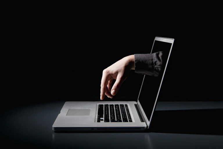 A hand reaching out of a laptop screen to type on a laptop