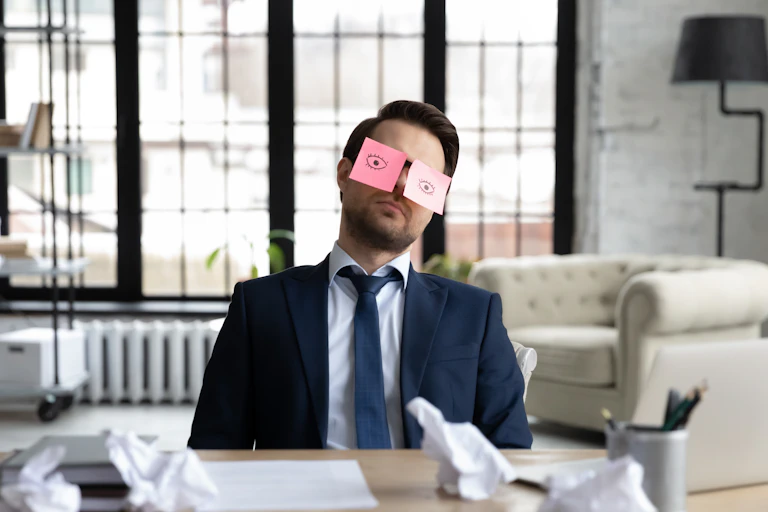 un homme à son bureau en train de faire la sieste, avec deux post it avec des yeux ouverts grossièrement dessinés pour faire croire qu'il travaille.