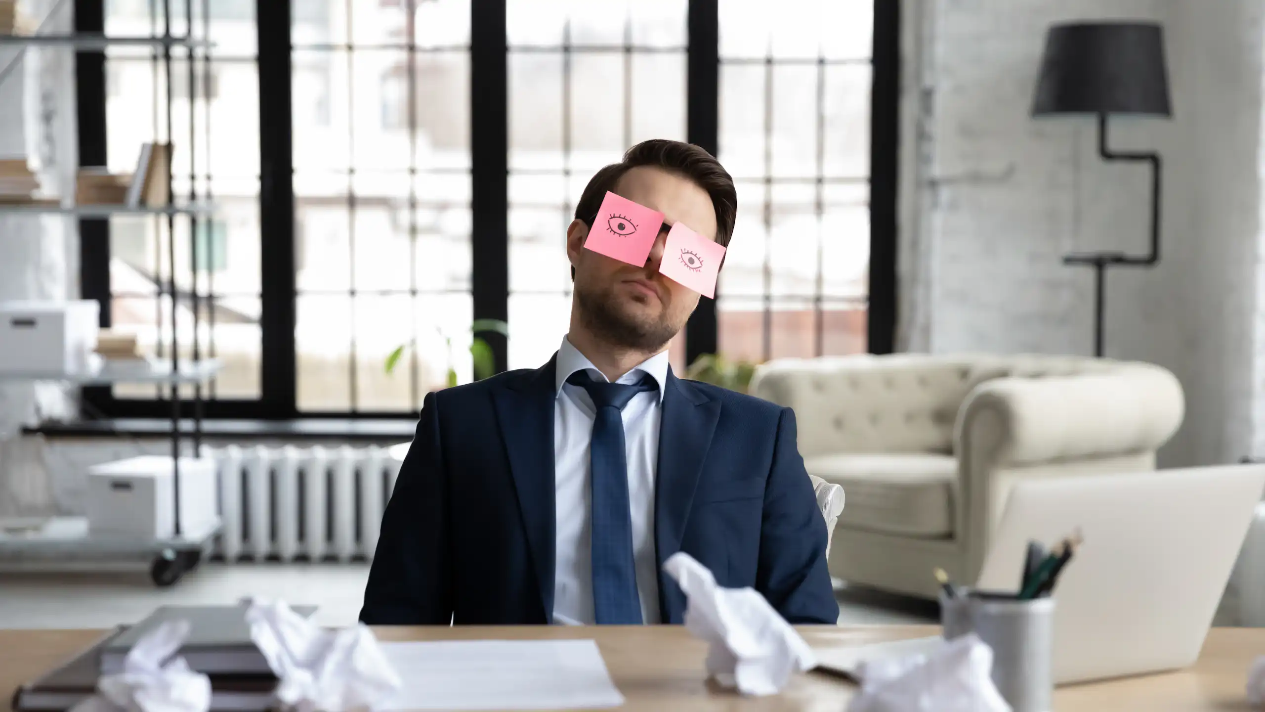 un homme à son bureau en train de faire la sieste, avec deux post it avec des yeux ouverts grossièrement dessinés pour faire croire qu'il travaille.