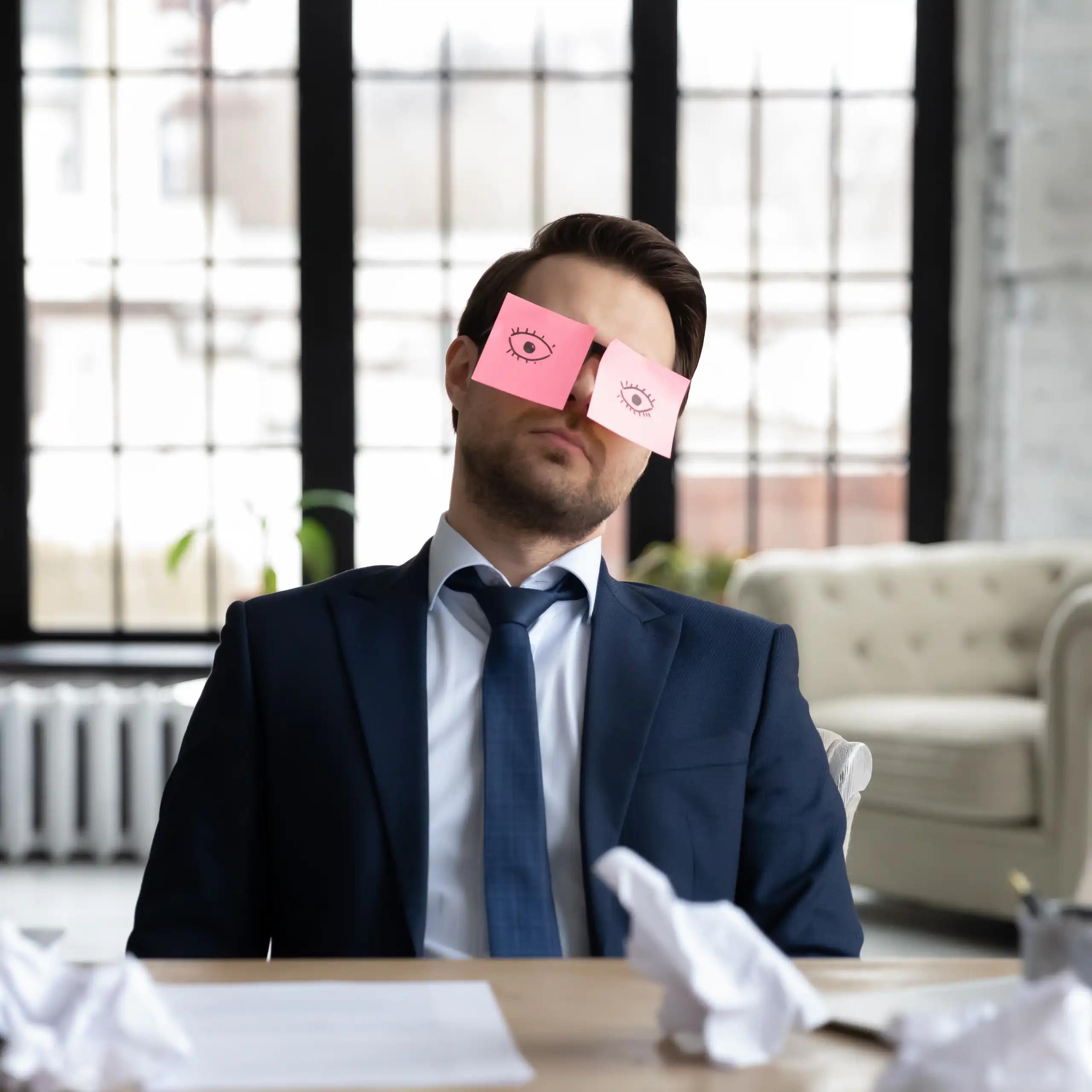 un homme à son bureau en train de faire la sieste, avec deux post it avec des yeux ouverts grossièrement dessinés pour faire croire qu'il travaille.