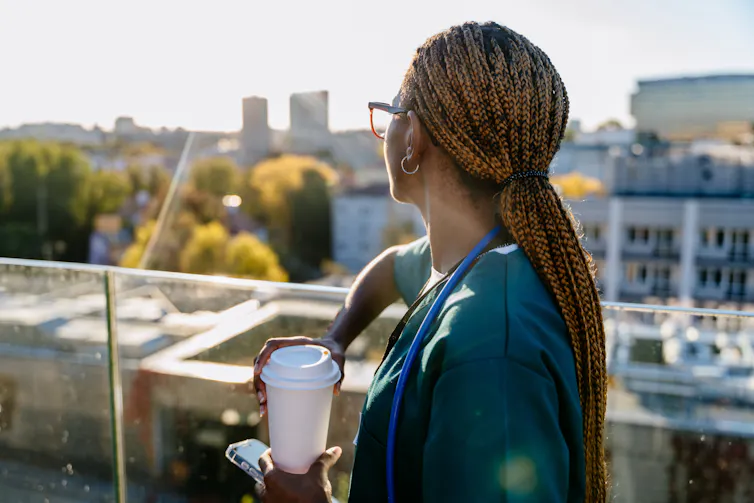 female healthcare worker on a break outside on a hospital balcony with a coffee and her phone in her hand.