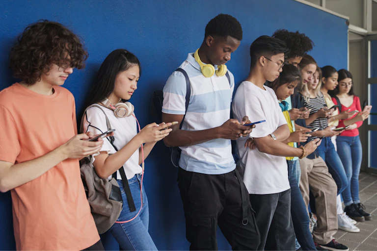 Un grupo de adolescentes de diferentes orígenes étnicos que utilizan sus teléfonos inteligentes frente a una pared azul