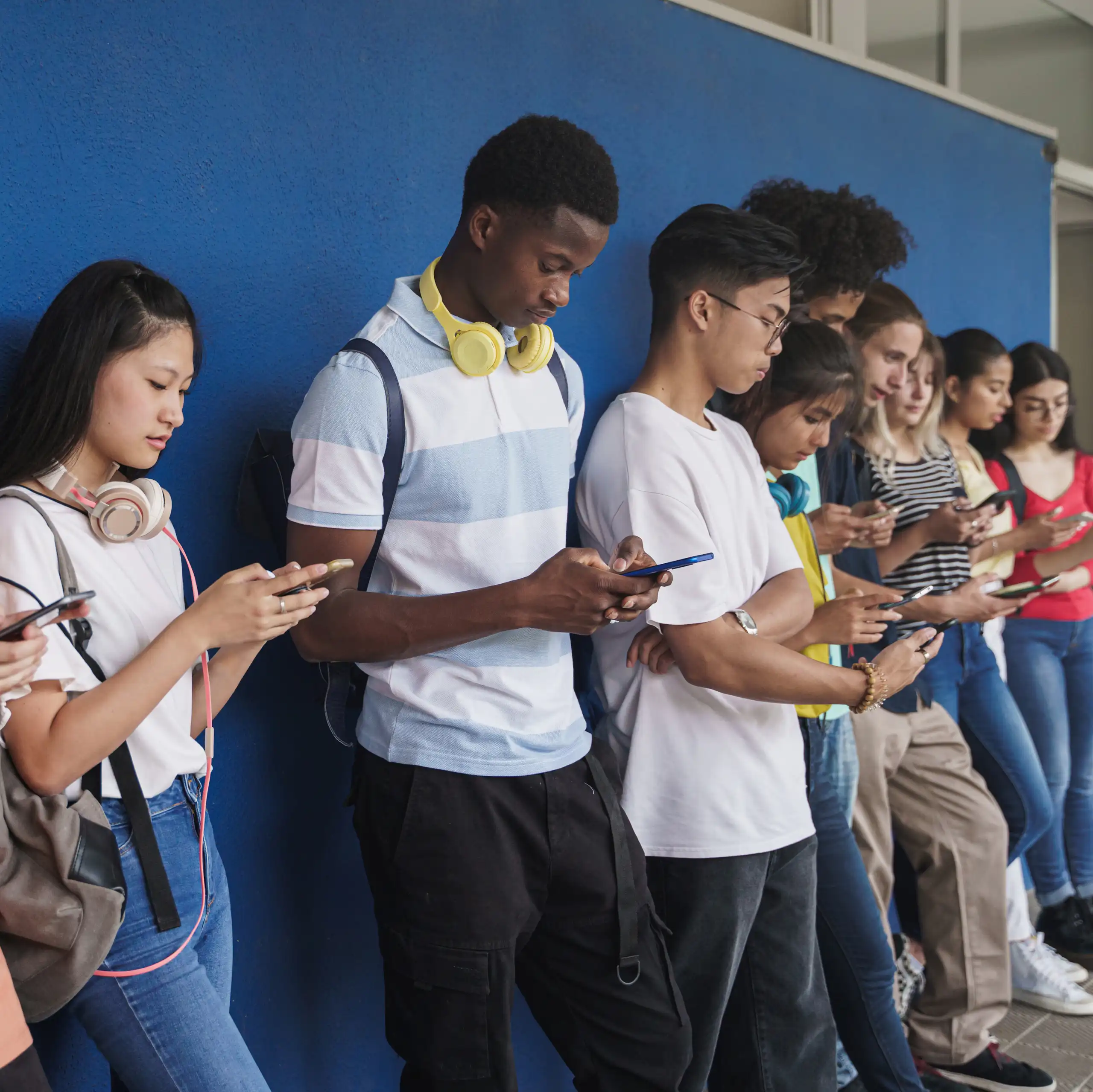 Un grupo de adolescentes de diferentes orígenes étnicos que utilizan sus teléfonos inteligentes frente a una pared azul