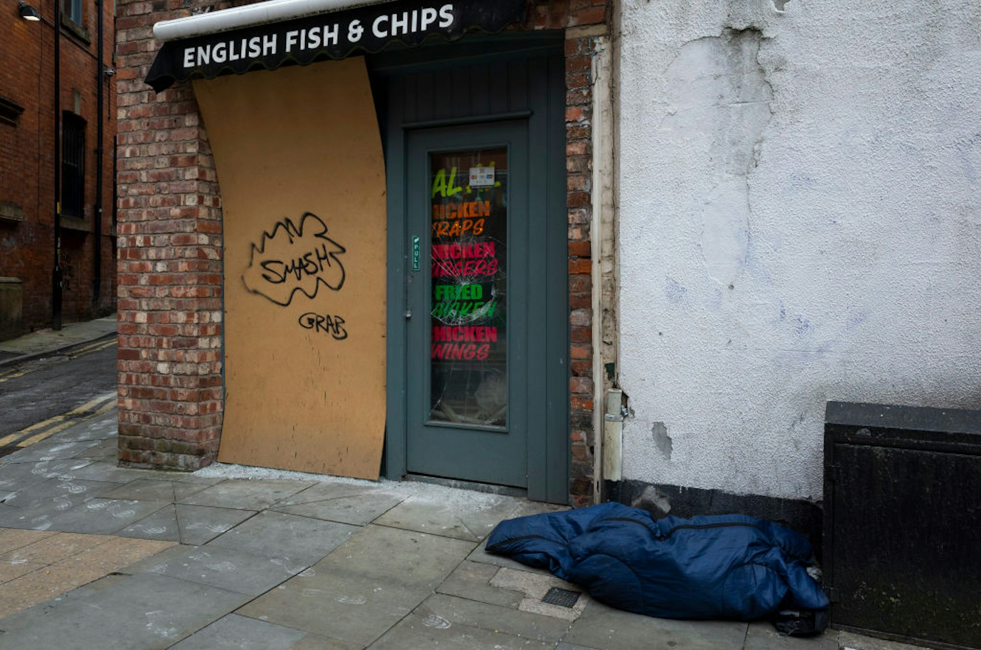 A person sleeps in a blue sleeping bag next to a boarded-up storefront.
