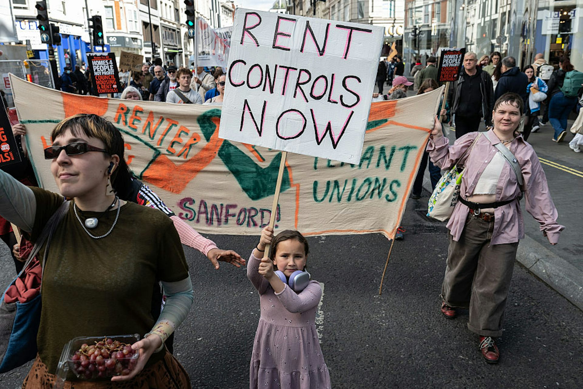 Young girl holds sign reading 'Rent Controls Now' as a large group of people march behind her while holding banners and signs.