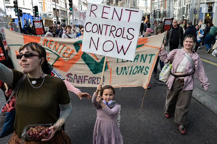 Young girl holds sign reading 'Rent Controls Now' as a large group of people march behind her while holding banners and signs.