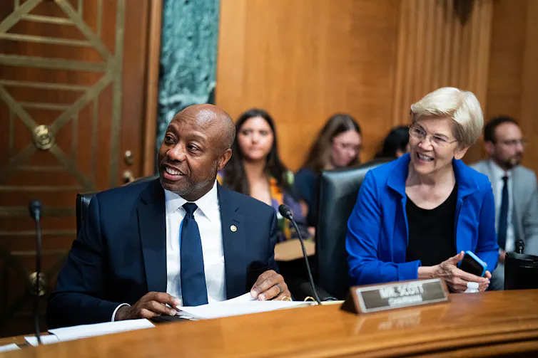 Middle-aged Black man wearing a suit and middle-aged white woman wearing a blue shirt smile while sitting at a dais.