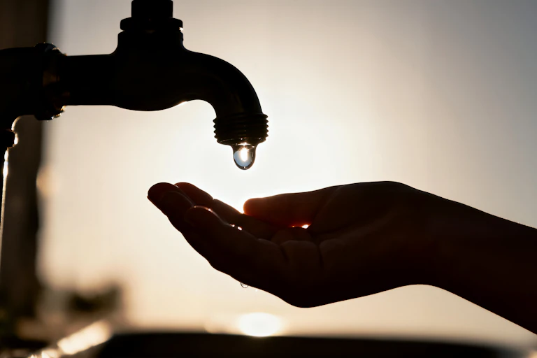 Una mano y un grifo a contraluz mientras cae una gota de agua.