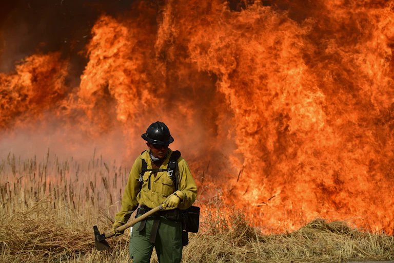 A man with an axe and a hard hat walks in front of bright orange red blazing fire.