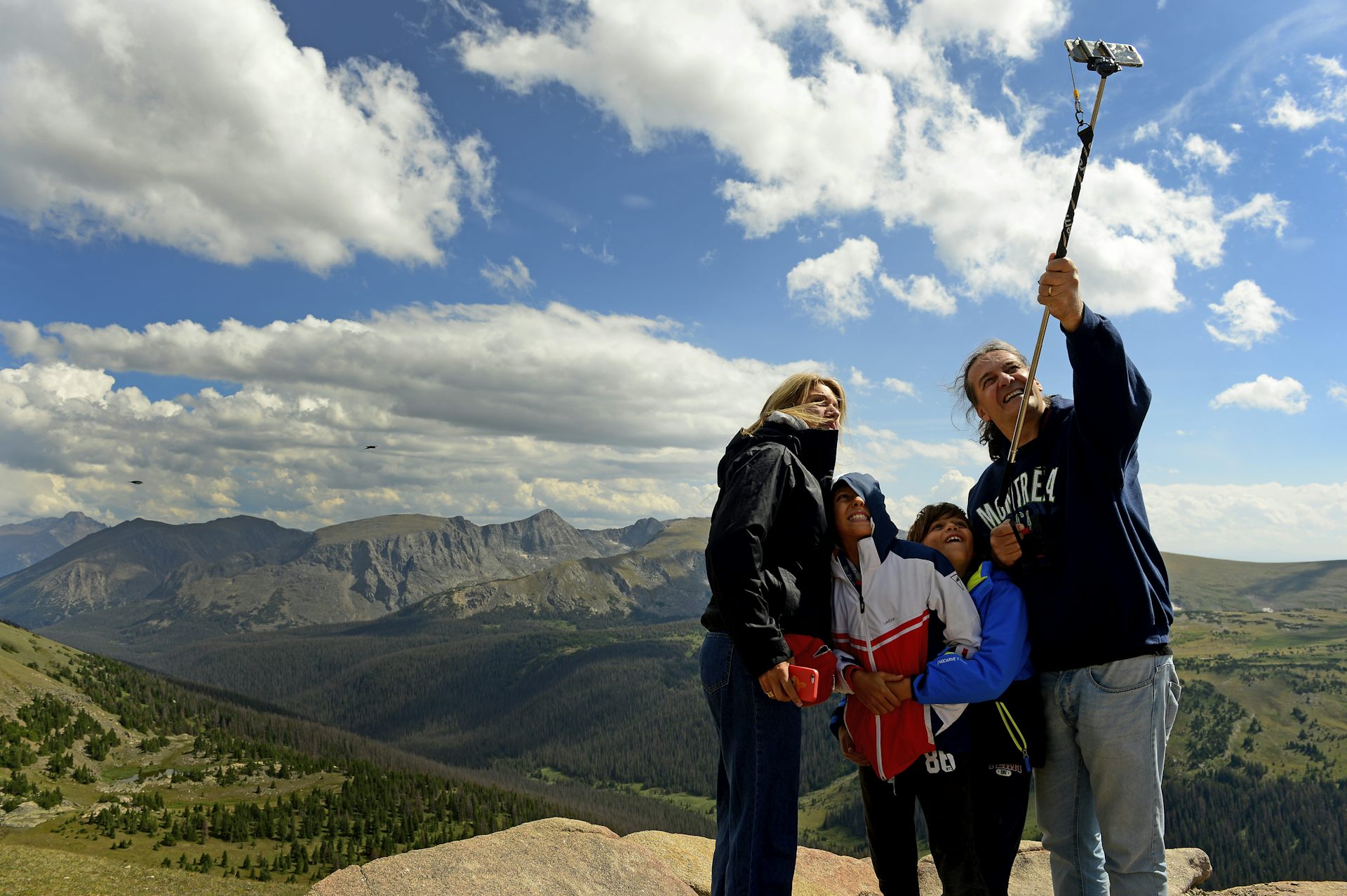Dos adultos y dos niños se reúnen frente a un palo para selfies con las montañas detrás.