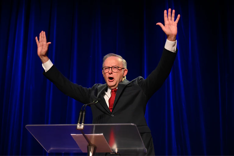 Anthony Albanese at a podium puts both hands up in the air