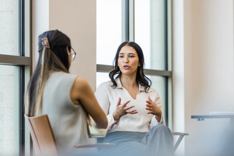 Two women sit on chairs and speak in a hall