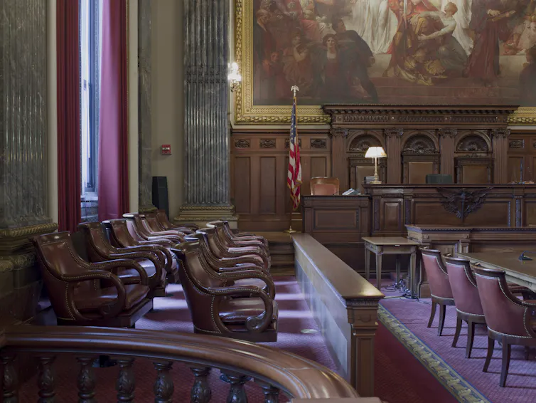 A photograph of an ornate, high-ceilinged room with heavy wooden chairs and a large mural on one wall.