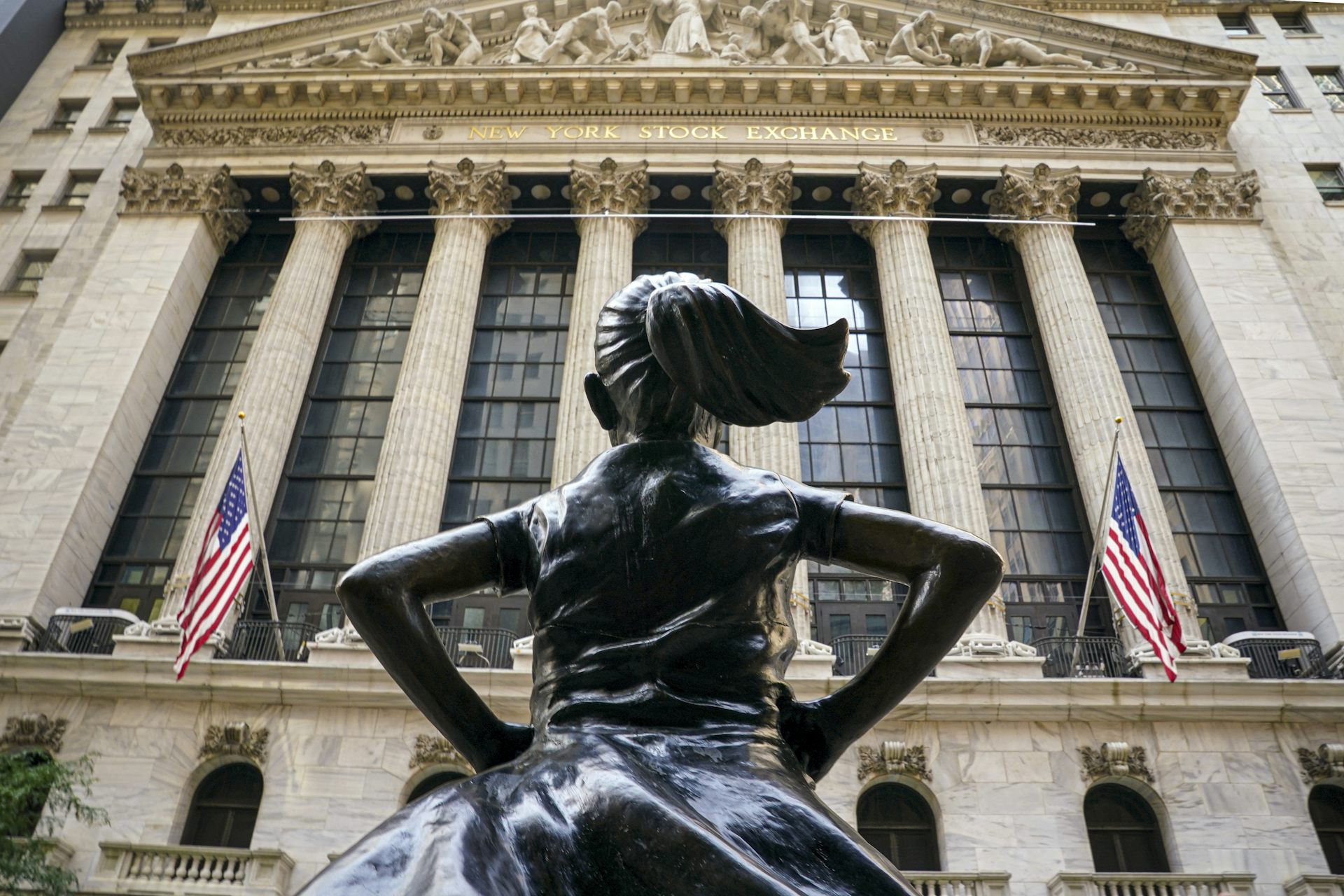 In front of a building with large pillars stands a statue of a girl with a ponytail, hands on her hips and seen from behind.
