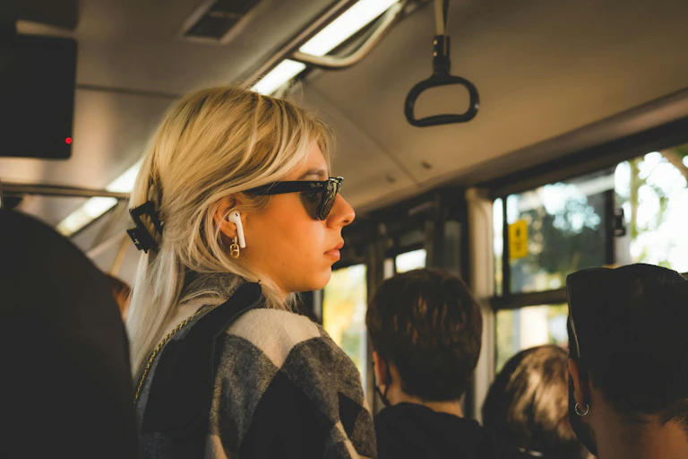 A woman with blonde hair commuting on a bus, listening to white wireless headphones.