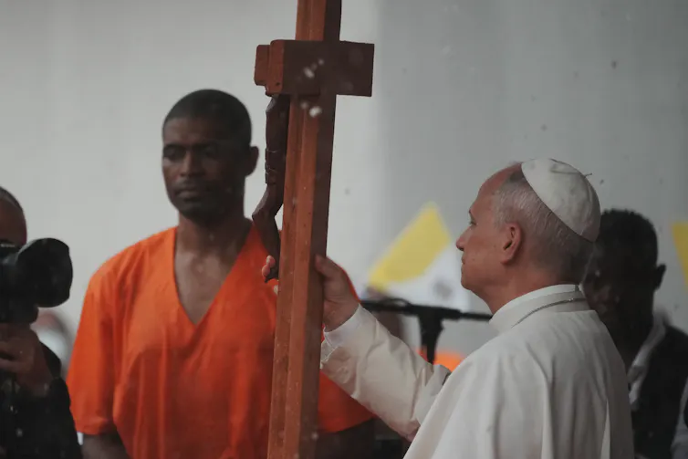 The pope holds a cross as he stands beside a man in a saffron top, while others look on.