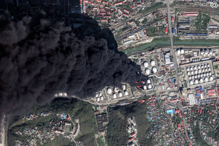 satellite image of an oil refinery on fire, thick column of black smoke.
