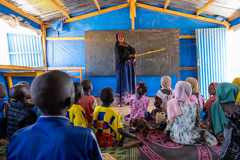 Woman stands at a blackboard in front of children seated on the floor