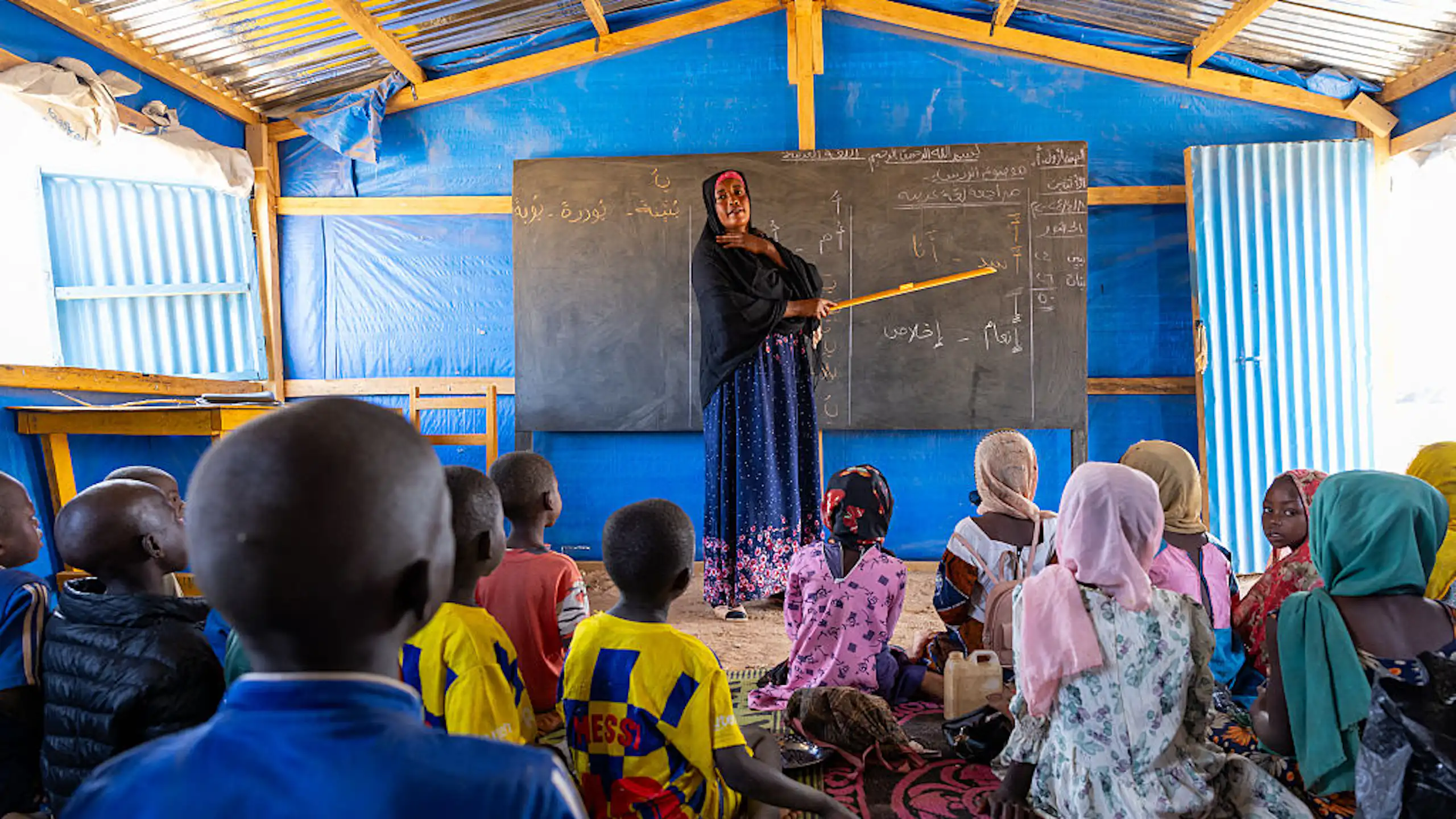 Woman stands at a blackboard in front of children seated on the floor