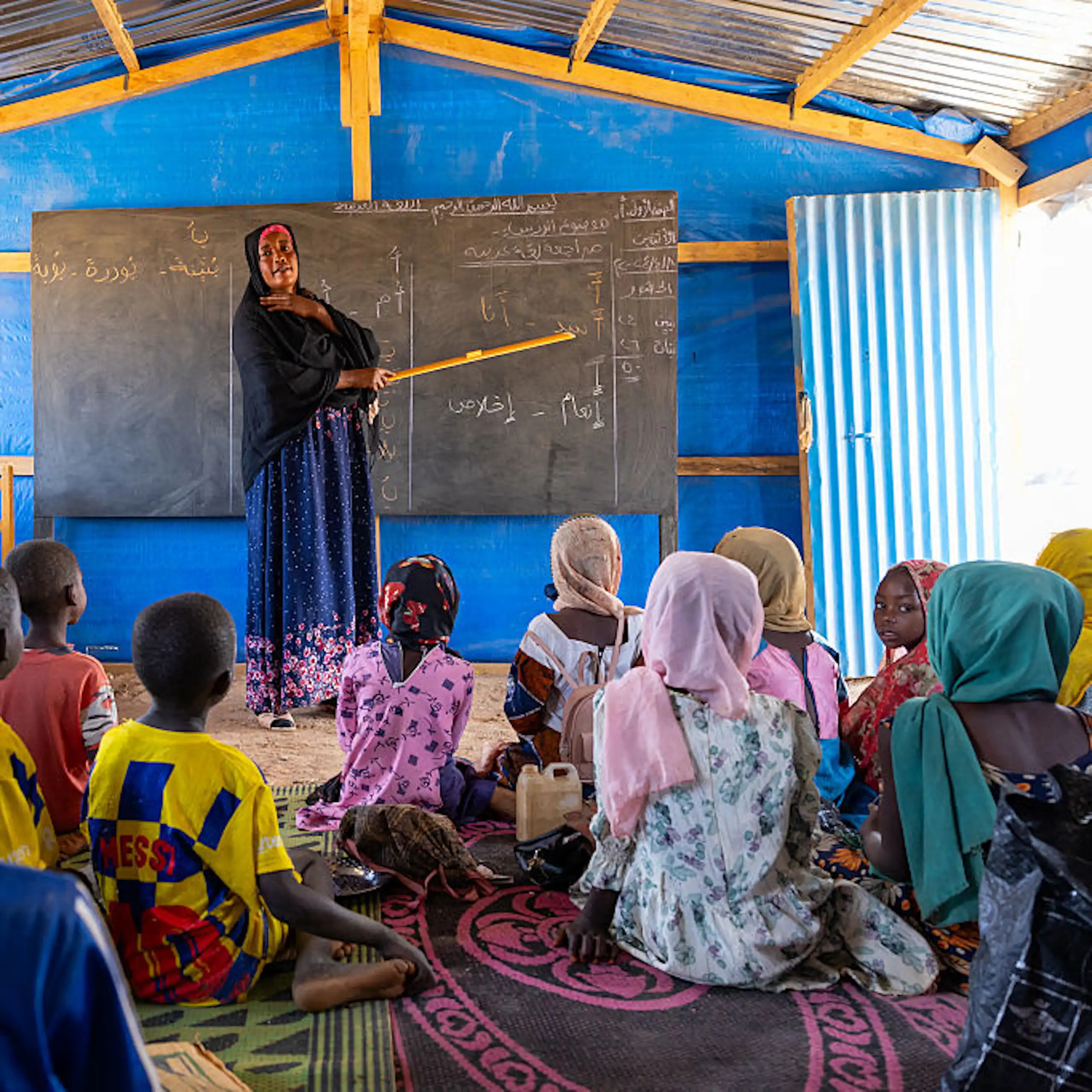 Woman stands at a blackboard in front of children seated on the floor