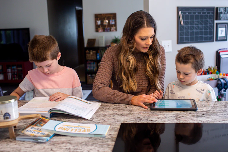 A woman with brown hair sits between two boys at a kitchen counter. They all look down at books and a tablet.