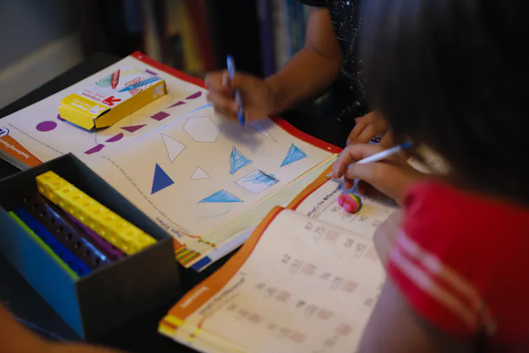 Two children, whose arms and shoulders are seen in this cropped photo, hold pens and lean over workbooks, one of which has photos of triangles on it.