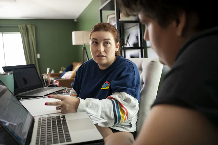 A woman wearing a long-sleeve shirt holds two fingers up near a laptop, as a teenage boy looks at the laptop and sits next to her.