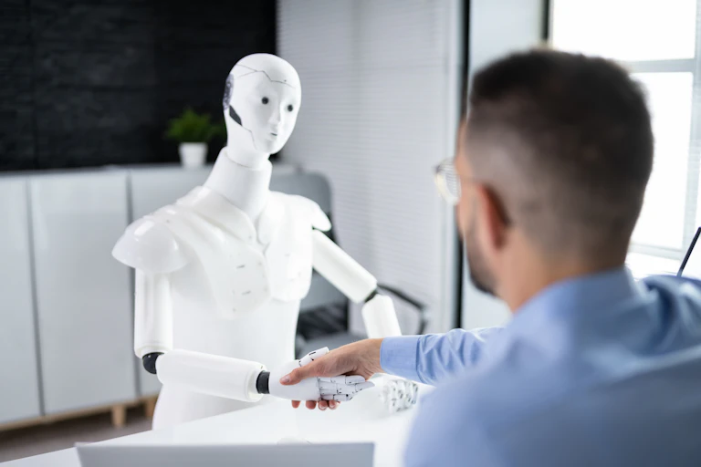 A man wearing glasses and blue work shirt, facing away, shakes hand of a white robot at a desk.