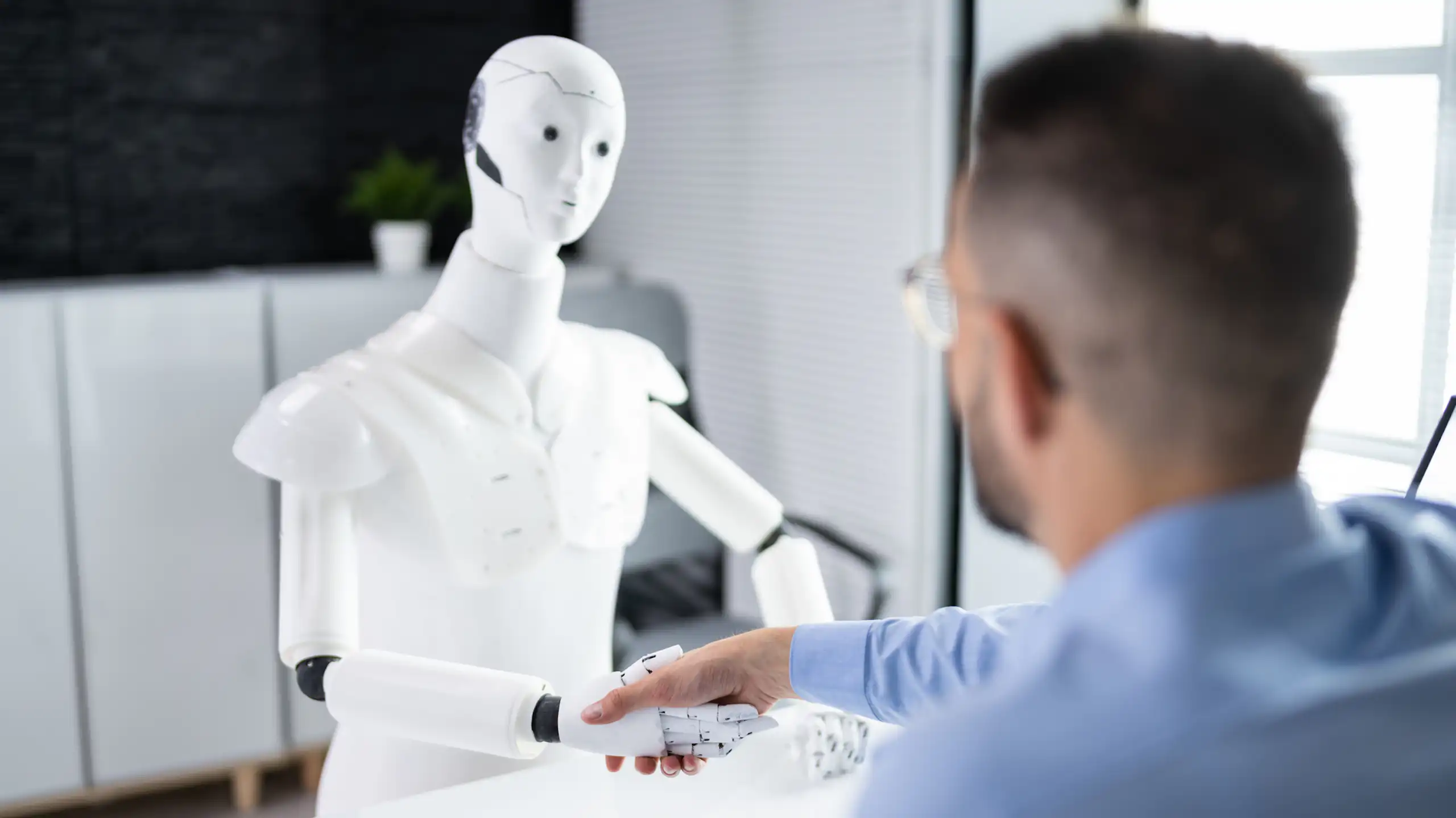 A man wearing glasses and blue work shirt, facing away, shakes hand of a white robot at a desk.