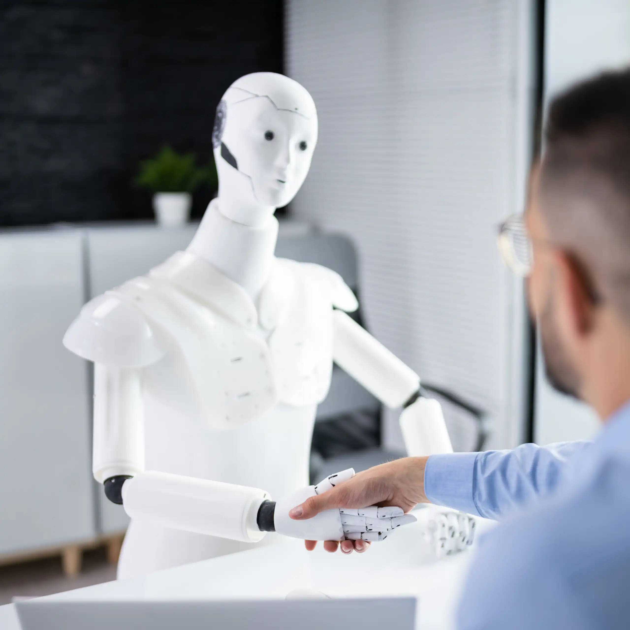 A man wearing glasses and blue work shirt, facing away, shakes hand of a white robot at a desk.