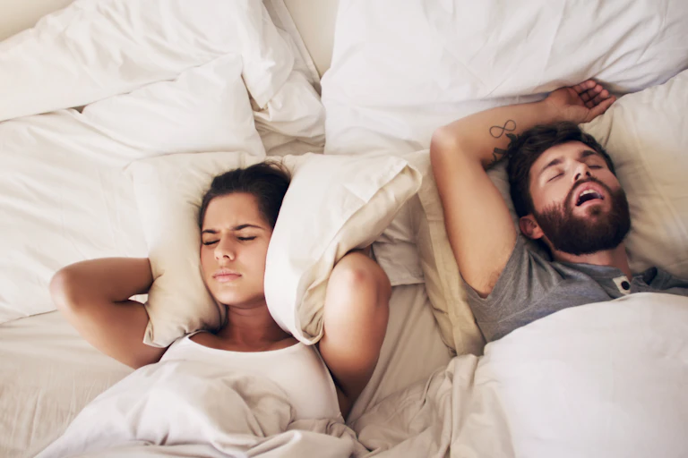 Down-facing view of a young woman covering her ears with a pillow while her partner snores in bed.