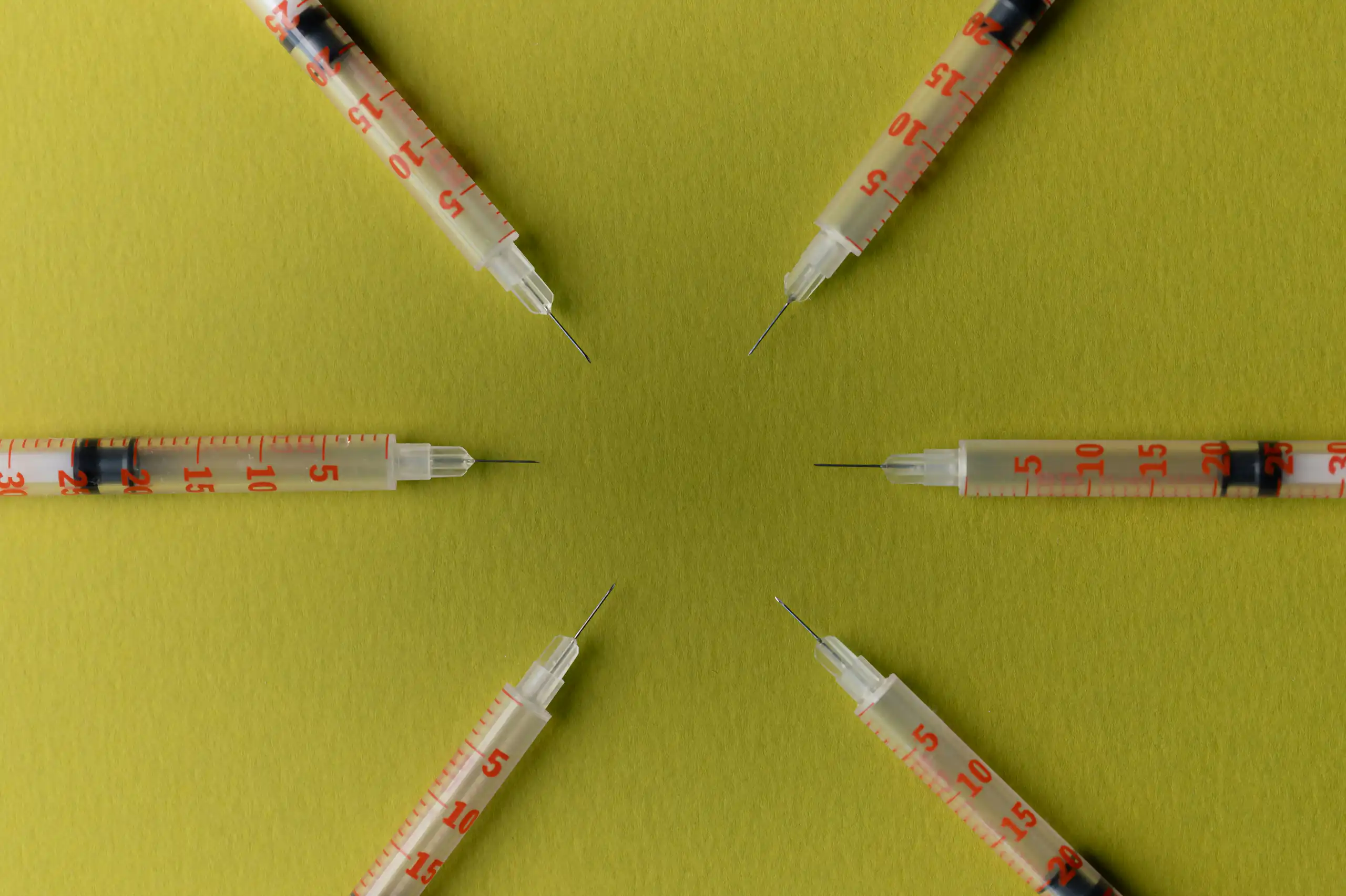 Six syringes arranged in a circle pointing inward against a yellow background