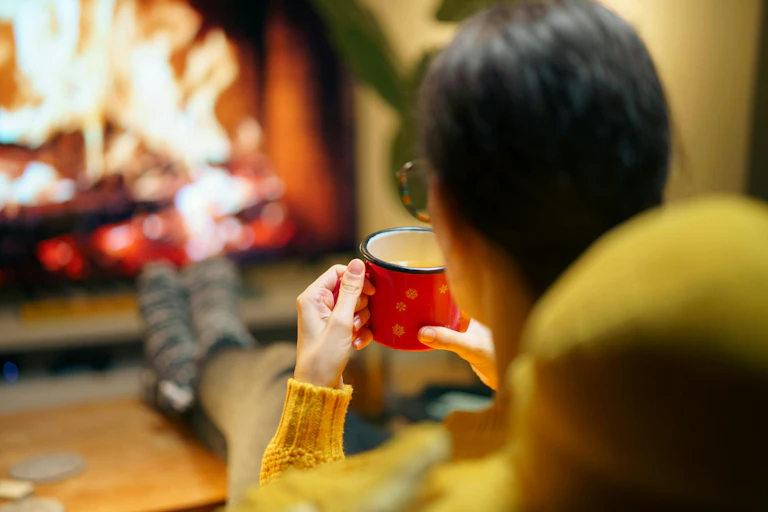 A woman sits holding a tea mug with her feet towards a fire.