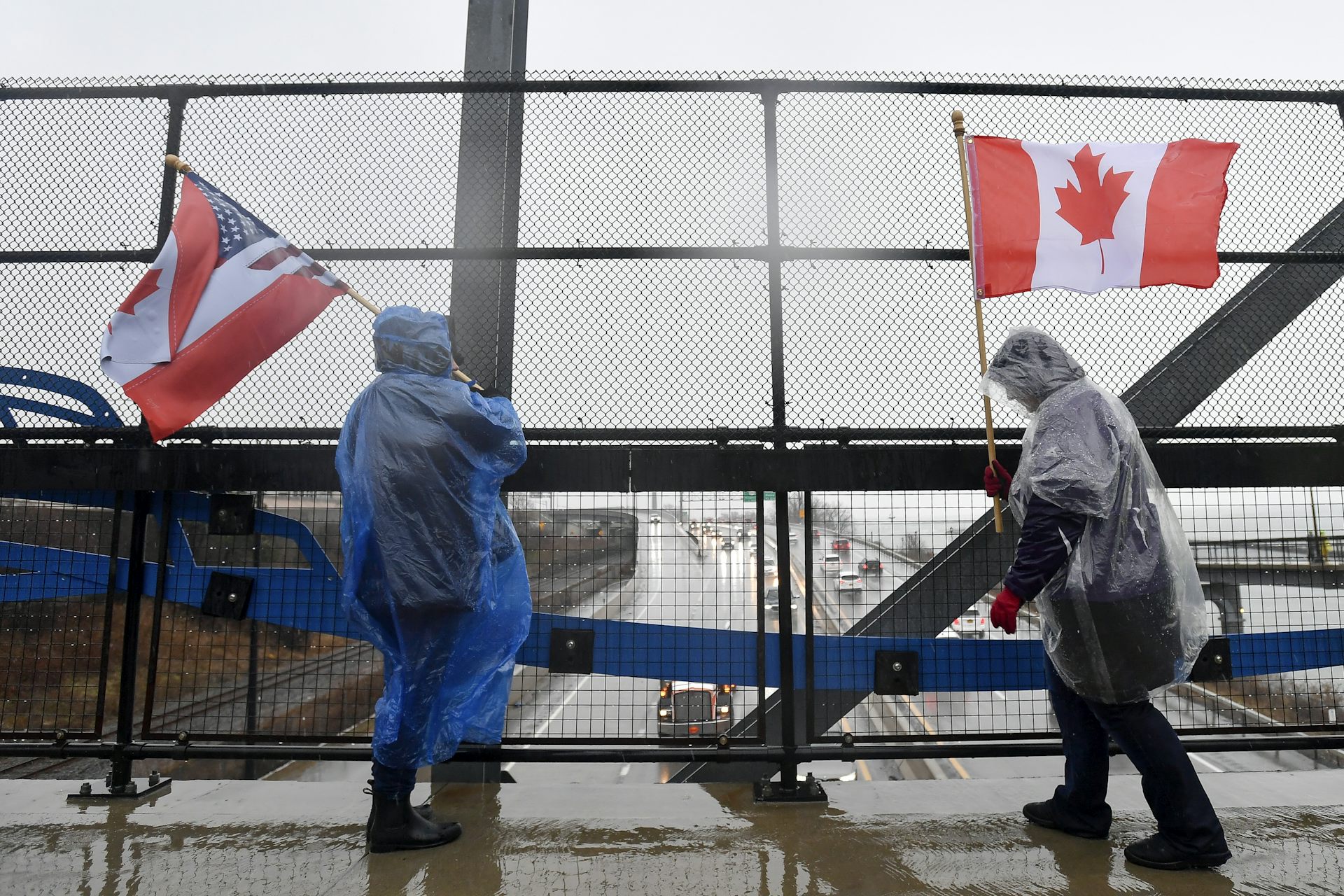 Two Canadian protesters wearing rain ponchos and carrying flags stand on the Peace Bridge border crossing in Buffalo, N.Y., on a gray and rainy day.