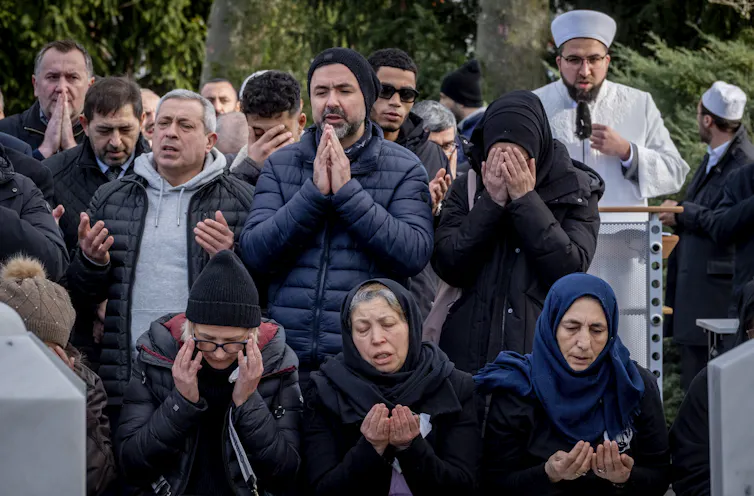 A group of mourners at a grave site have their hands up in prayer while some are crying.