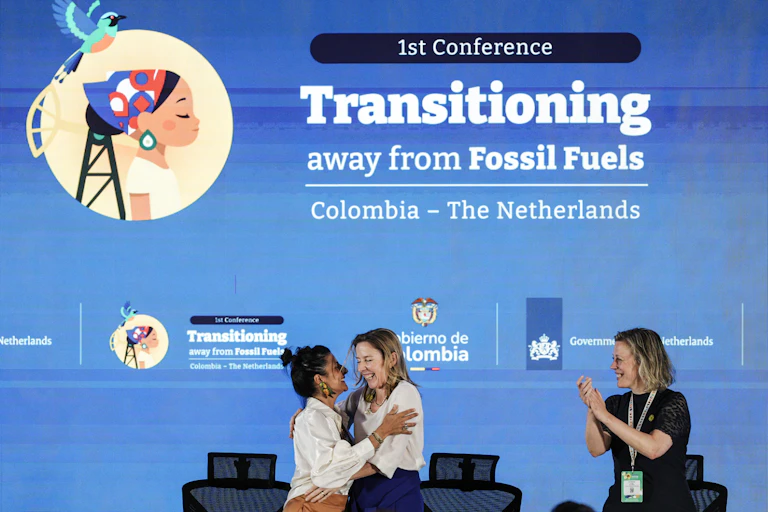 two women on a stage hug as a third claps, behind them is a large screen with the words: 1st conference transitioning away from fossil fuels