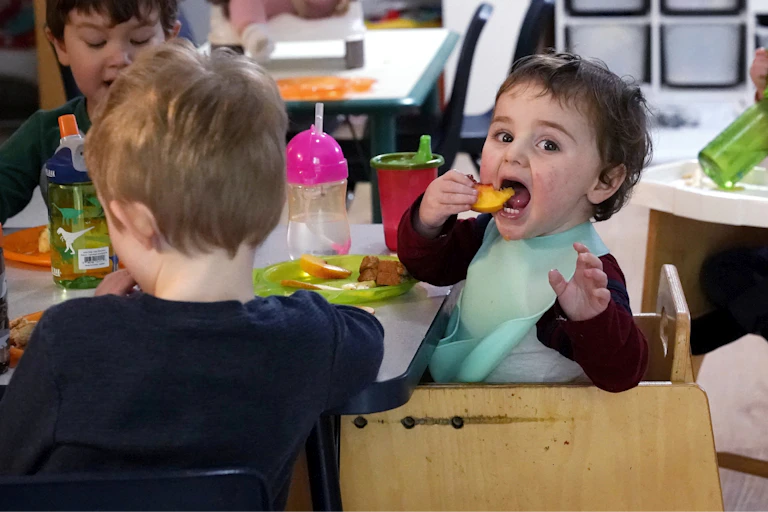 A toddler in a high chair has a huge open mouth and a hand raised and looks content feeding themselves a piece of fruit.
