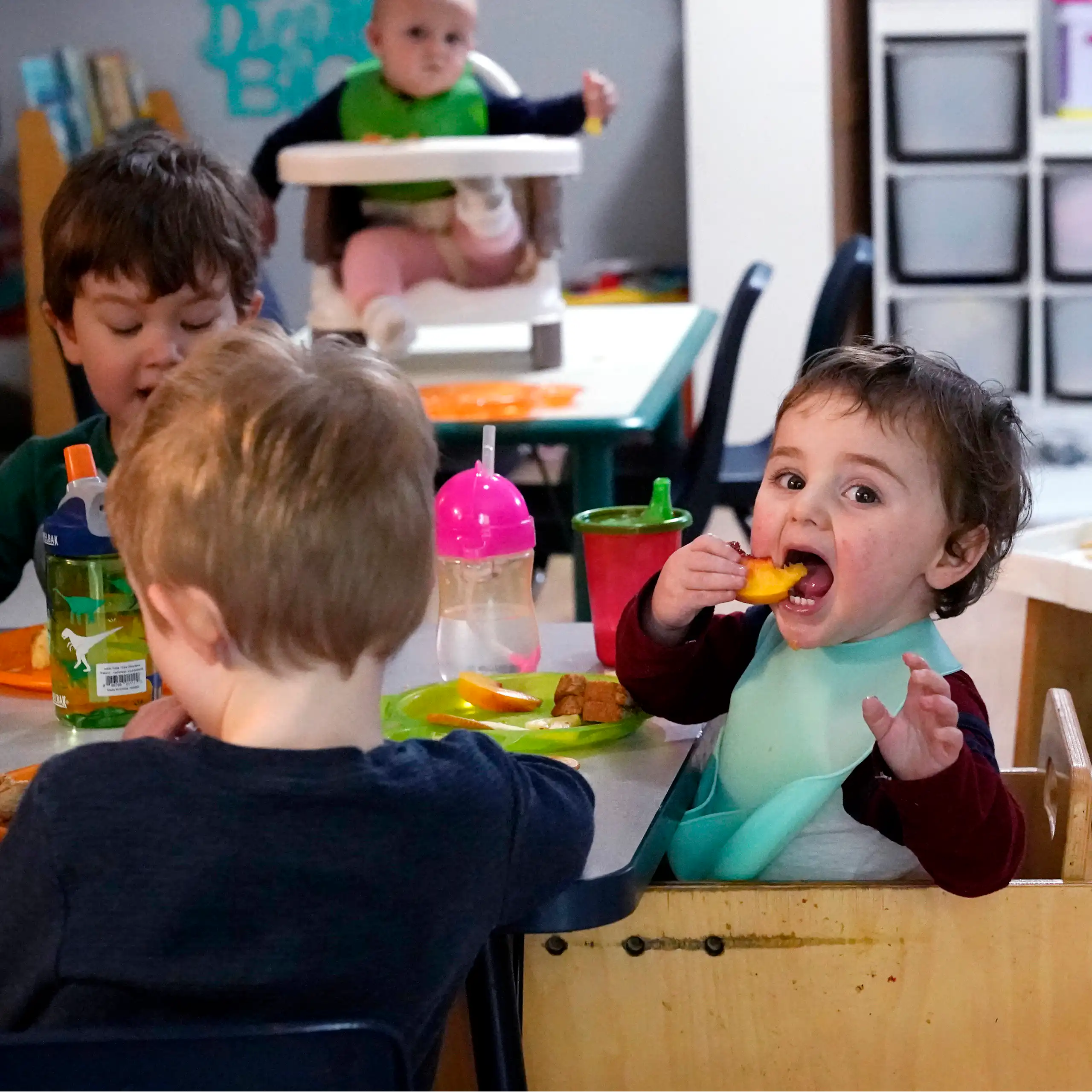 A toddler in a high chair has a huge open mouth and a hand raised and looks content feeding themselves a piece of fruit.