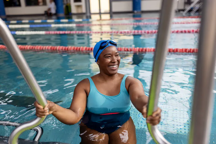 Smiling Black woman in swimsuit holding onto rails in indoor pool.
