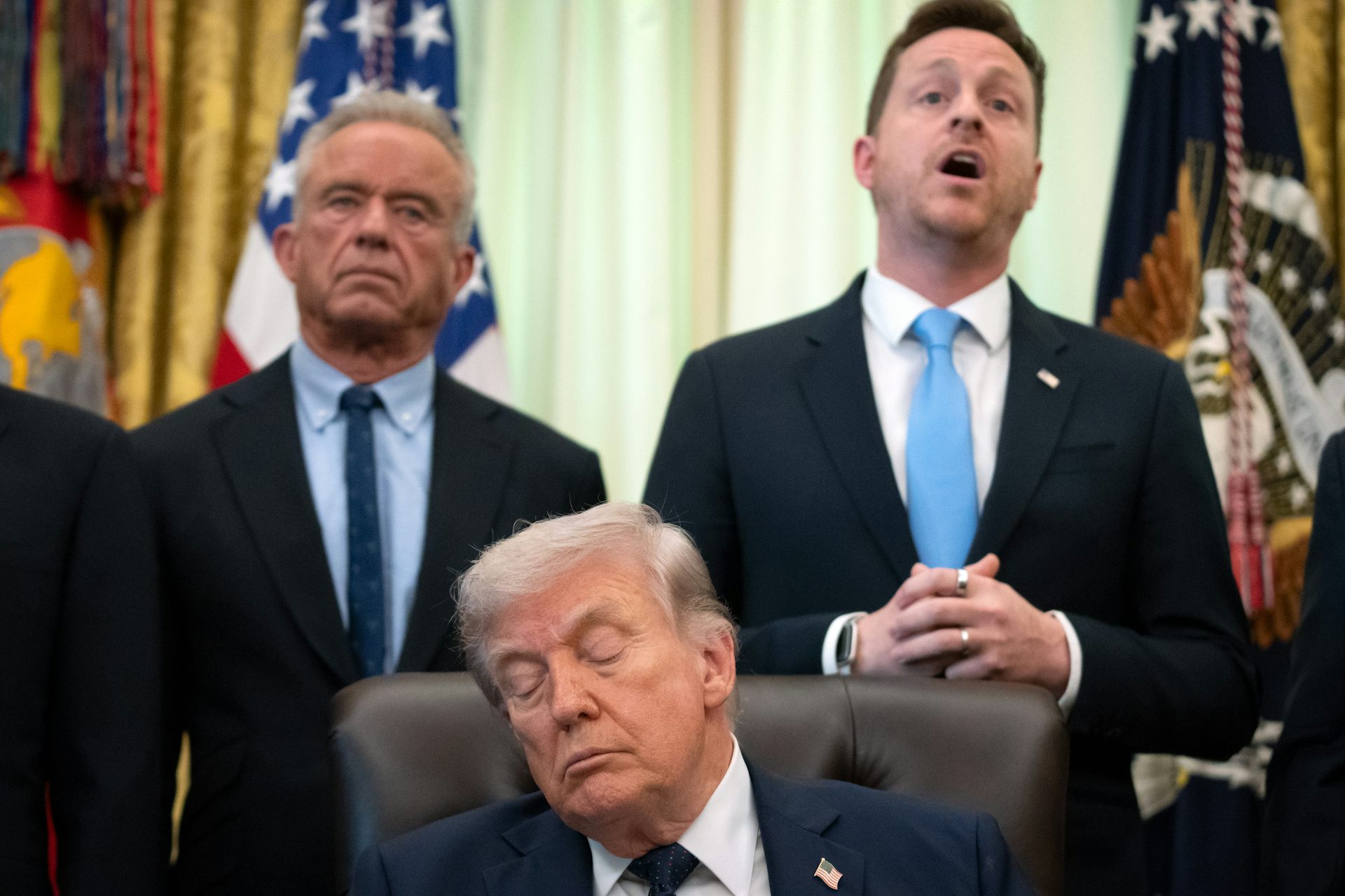 An older man with orange-hued skin snoozes in an office chair with two men standing behind him.