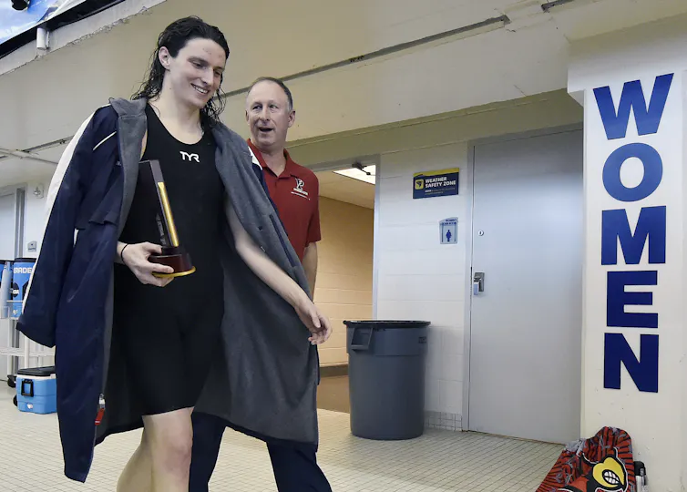 A woman with dark hair and a long jacket smiles and holds a trophy, walking next to a man in front of a woman's bathroom sing.