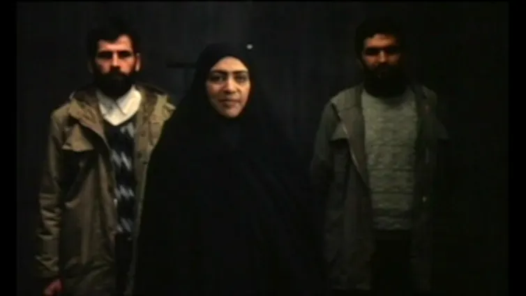 A women in a black hijab and abaya (robe) stand straight, with two men behind her, also facing the camera.