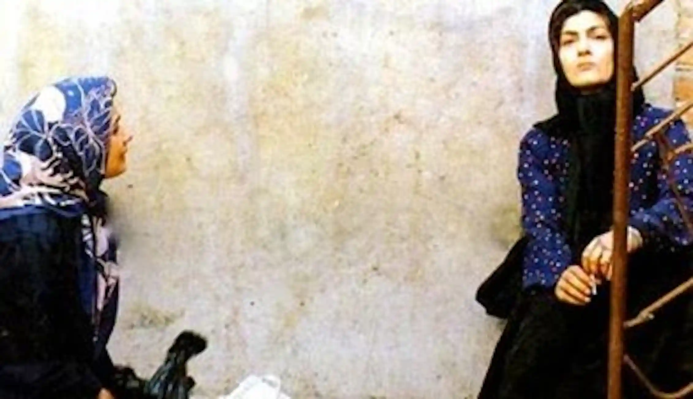 A woman in a black hijab sits at the end of a staircase, while another women to the left, in a blue patterned hijab, speaks to her.
